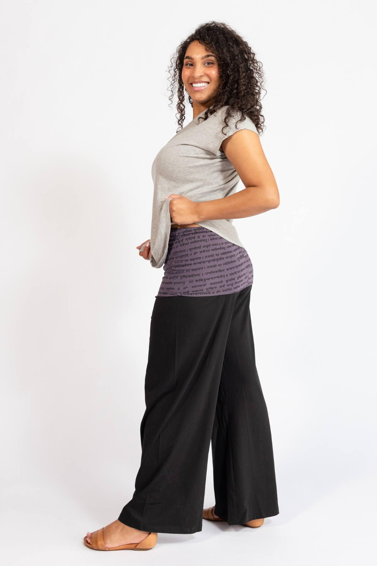 Woman wearing black turn over waist pants, turned to the left and against a white background