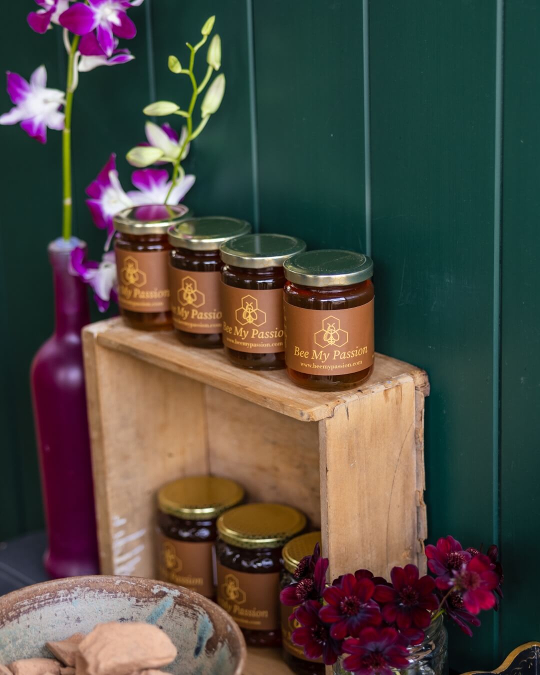 Four glass jars of local honey on top of a rustic timber shelf. Small flowers surround.