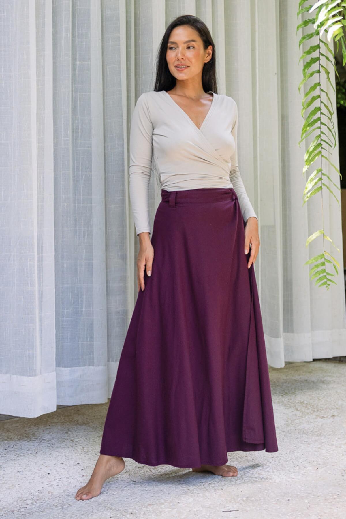 Woman standing in front of a breezy white curtain, wearing a long purple wrap skirt and white wrap around top.