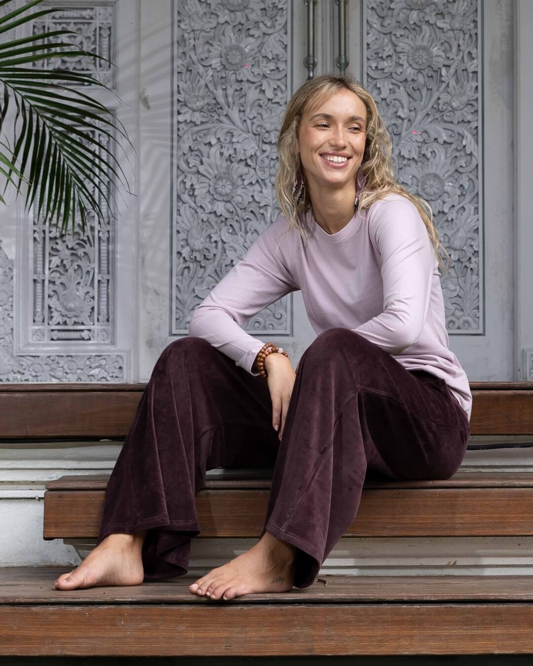 Woman is sitting relaxed on the edge of timber stairs. She is wearing a cocoa coloured pair of velvet pants and a blush pink long sleeve organic cotton top. 
