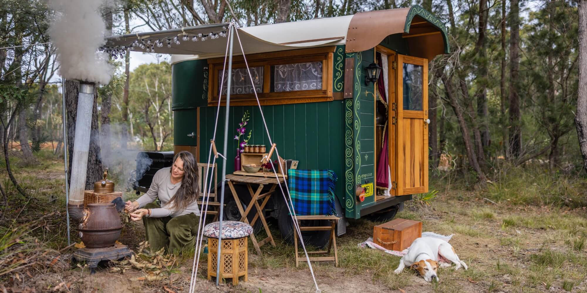 A Woman sitting near a lighted stove under an awning in the Australian bush. Her dog sits beside the door of her gypsy wagon. She is wearing velvet pants.