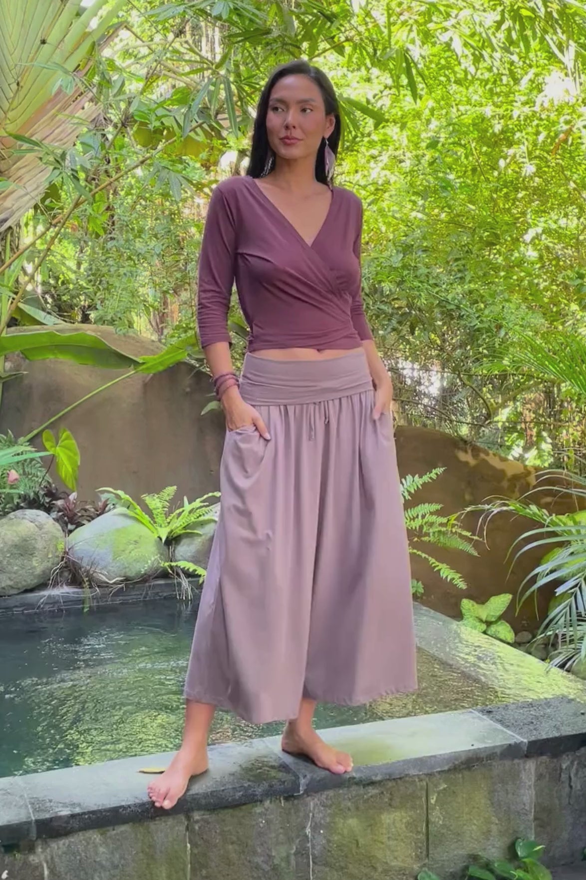 Woman wearing an organic cotton wide leg pants and wrap top outfit, balancing on the edge of a pool surrounded by jungle.